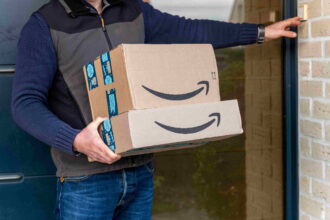 A person in a jacket holds two Amazon boxes, one on top of the other, while ringing a doorbell. The scene suggests a package delivery.