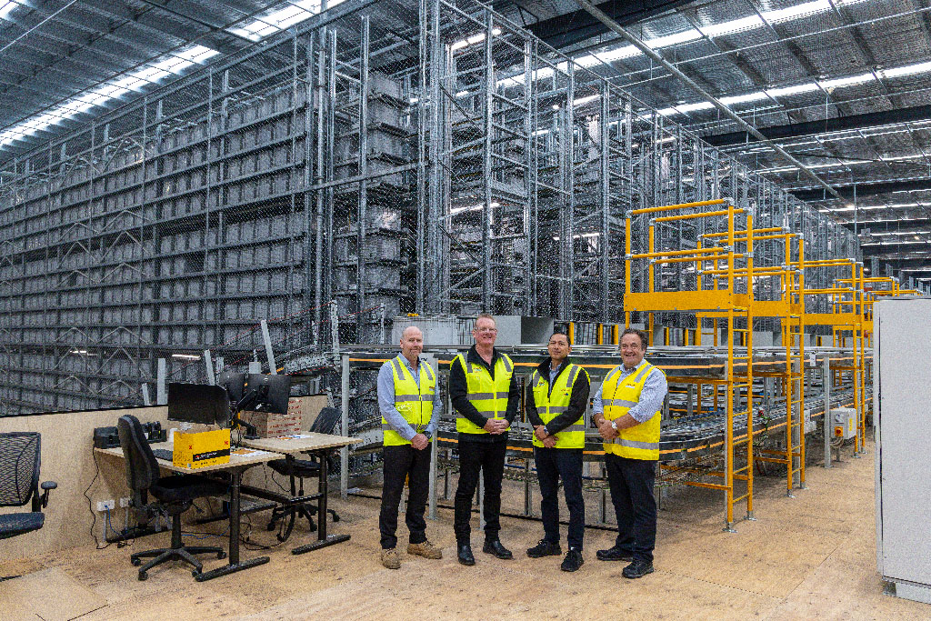 Four people in high-visibility vests stand in a modern warehouse, surrounded by towering metal shelves and yellow safety railings, conveying professionalism.