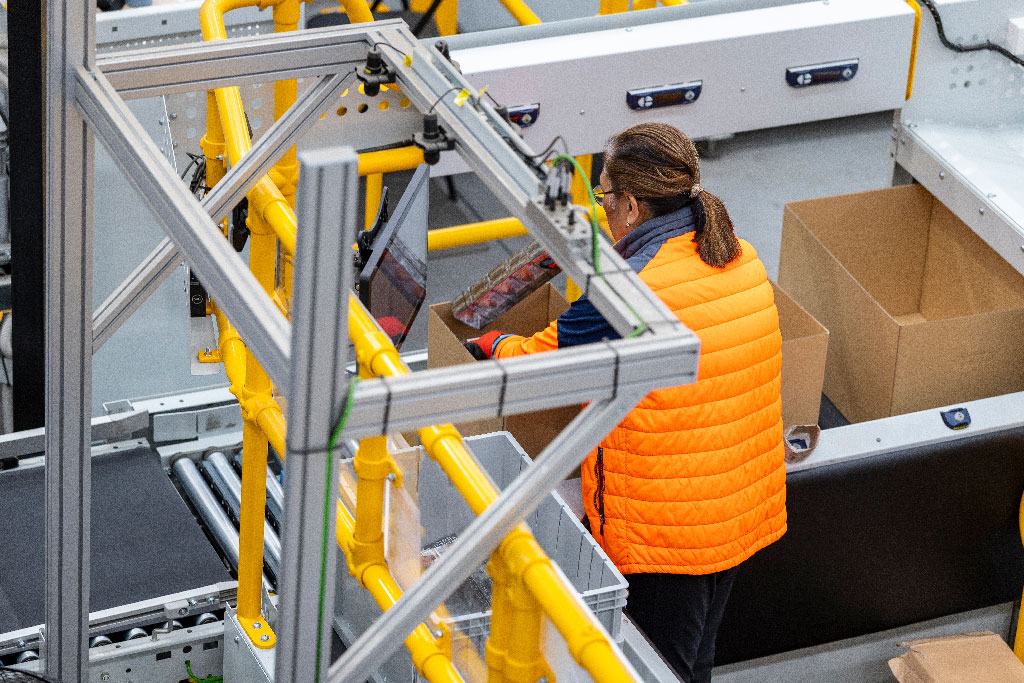 A person in an orange jacket works at a conveyor system in a factory, placing items into a cardboard box. The scene conveys efficiency and focus.