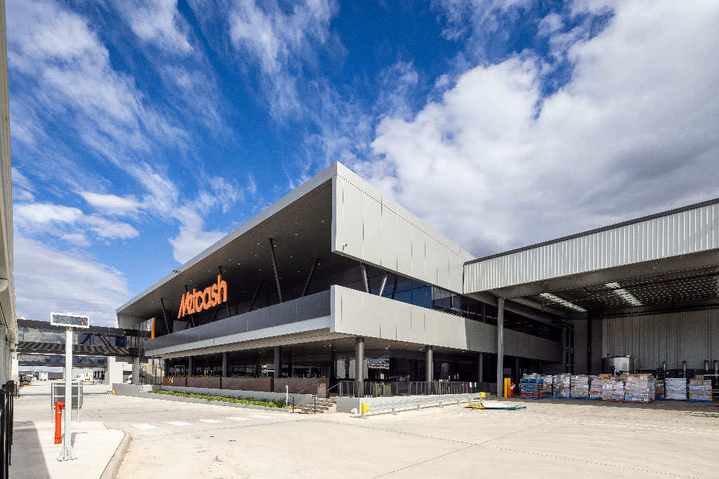 Modern warehouse exterior with "Metcash" logo, featuring sleek, angular architecture. Bright blue sky with clouds enhances the industrial setting.