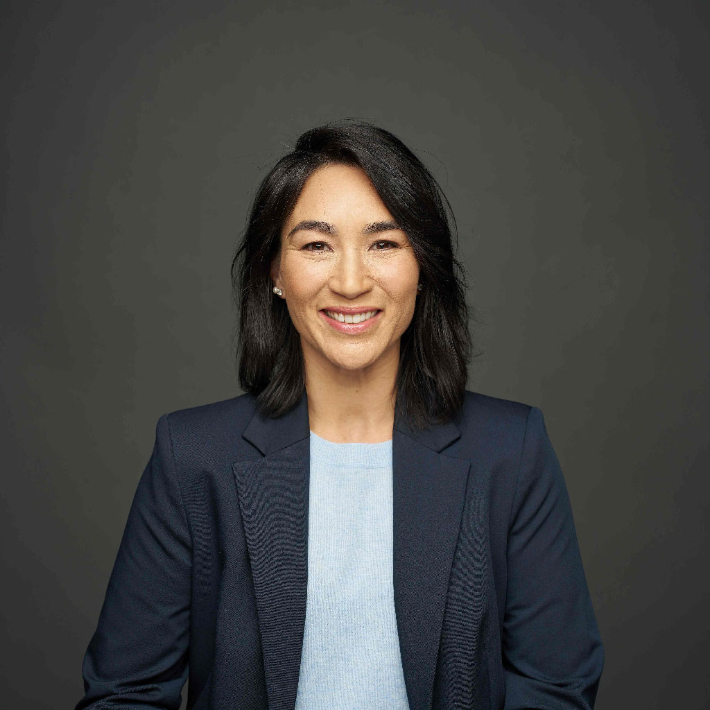 Smiling woman with shoulder-length black hair wearing a light blue blouse and navy blazer against a dark gray background, conveying confidence.