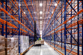 A vast, organized warehouse aisle with high, empty blue and orange racks. A worker on a forklift operates in the center, surrounded by pallets. Bright lighting.