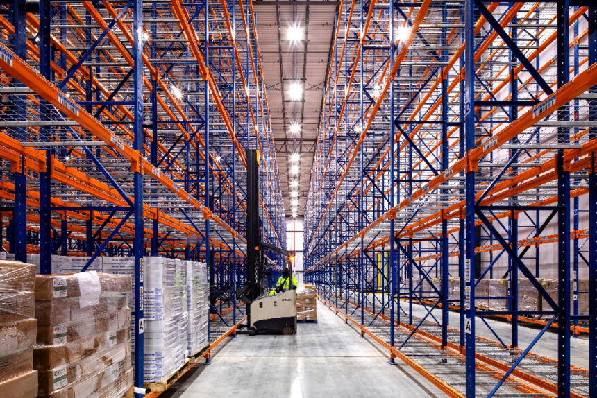 A vast, organized warehouse aisle with high, empty blue and orange racks. A worker on a forklift operates in the center, surrounded by pallets. Bright lighting.