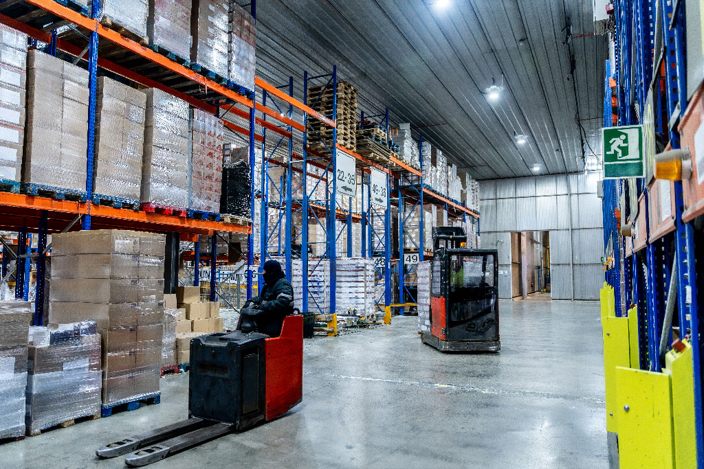 A forklift operator maneuvers through a large warehouse filled with stacked pallets and boxes. The scene is industrial, organized, and brightly lit.