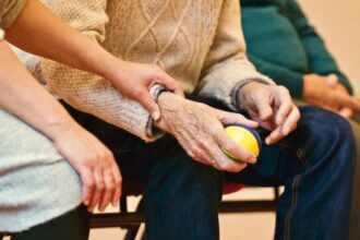 A person gently holds the wrist of an older adult seated in a chair, who holds a colorful ball. The scene conveys care and support.