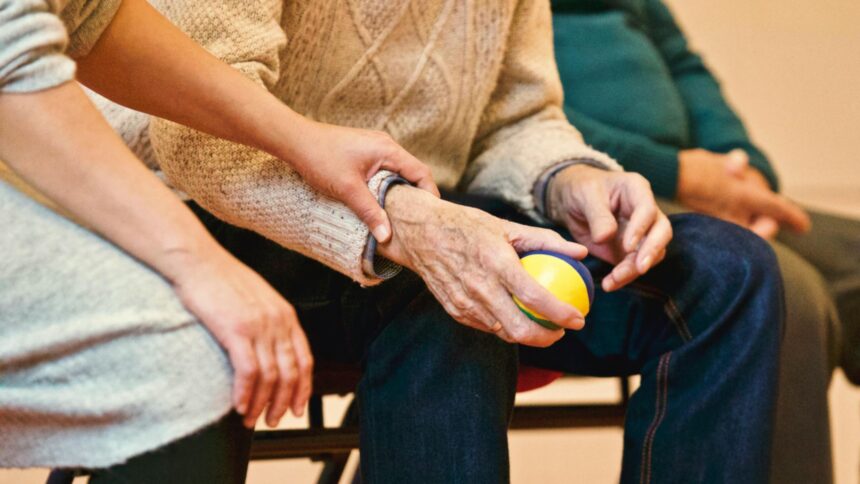 A person gently holds the wrist of an older adult seated in a chair, who holds a colorful ball. The scene conveys care and support.