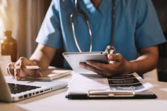 A healthcare professional in blue scrubs uses a tablet and laptop at a well-lit desk with medical documents. The scene conveys focus and efficiency.