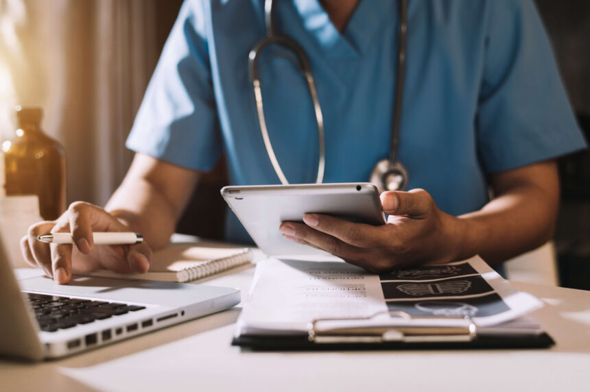 A healthcare professional in blue scrubs uses a tablet and laptop at a well-lit desk with medical documents. The scene conveys focus and efficiency.