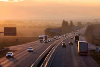 Busy highway at sunset with cars and trucks heading in both directions. The sky glows orange, casting a warm hue over the landscape and road.