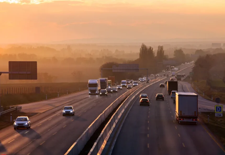 Busy highway at sunset with cars and trucks heading in both directions. The sky glows orange, casting a warm hue over the landscape and road.