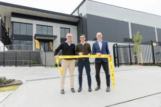 Three men stand outside a modern industrial building, smiling and holding a yellow ribbon with DHL branding, conveying a sense of celebration and achievement.