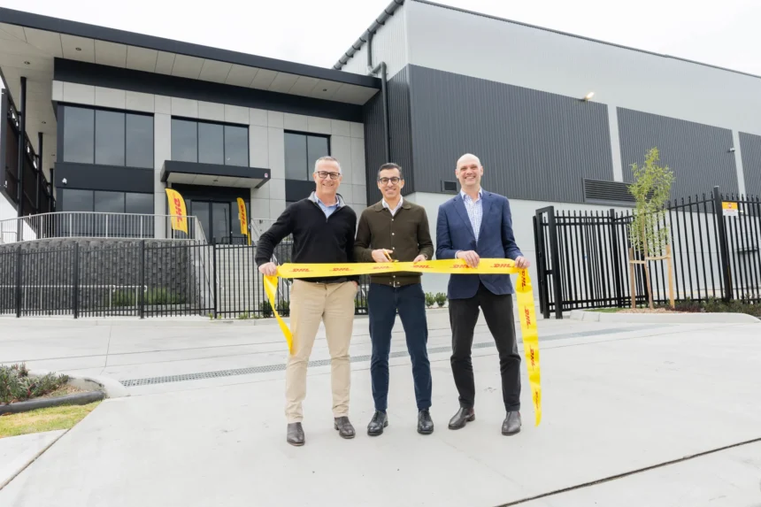 Three men stand outside a modern industrial building, smiling and holding a yellow ribbon with DHL branding, conveying a sense of celebration and achievement.