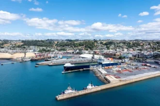 Aerial view of a bustling port with a large cargo ship labeled "Strait Link" docked. Surrounding the harbour are cranes, warehouses and distant hills under a blue sky.