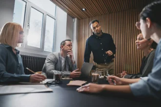 A diverse group of five professionals discusses around a modern conference table, with attentive expressions. The setting is a well-lit meeting room.