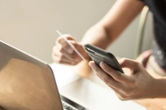 A person uses a smartphone with one hand and holds a credit card in the other, beside an open laptop, suggesting online shopping or banking.