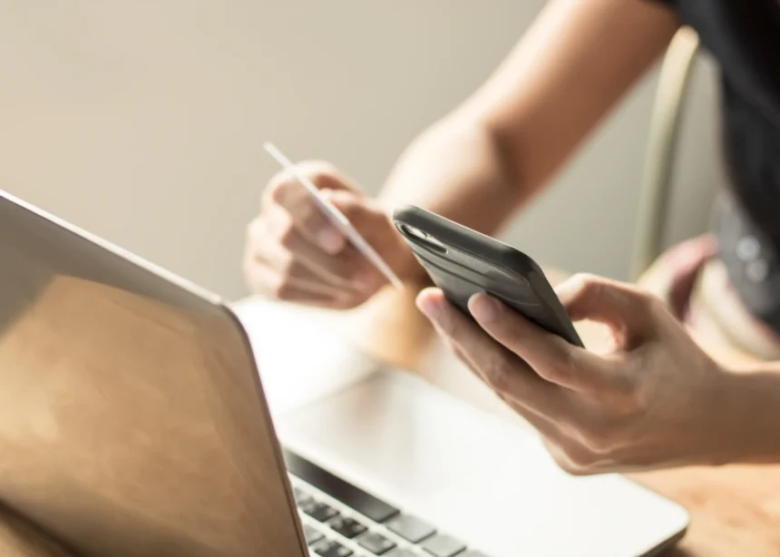 A person uses a smartphone with one hand and holds a credit card in the other, beside an open laptop, suggesting online shopping or banking.