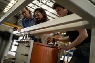 Three people examine machinery in a workshop, looking focused and engaged. The environment is industrial with visible structural beams.