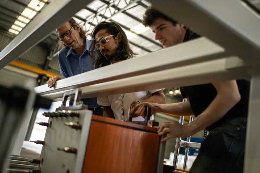 Three people examine machinery in a workshop, looking focused and engaged. The environment is industrial with visible structural beams.