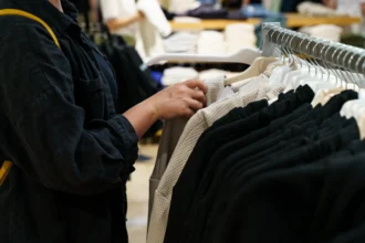 A person browses through jackets on hangers in a clothing store, with a focus on black and beige items. The setting feels casual and focused.