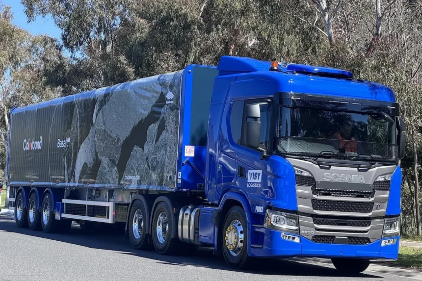 A blue Scania truck with a long trailer displaying a monochrome rock pattern and "Colorbond Basalt." It’s parked on a sunny road with trees surrounding it.