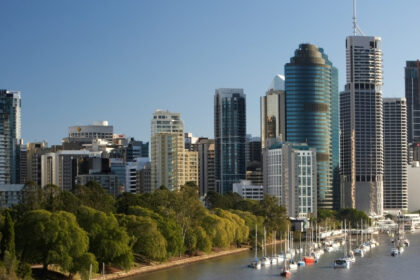 Brisbane city skyline viewed from the river, with high-rise buildings, parkland and moored boats along the waterfront.