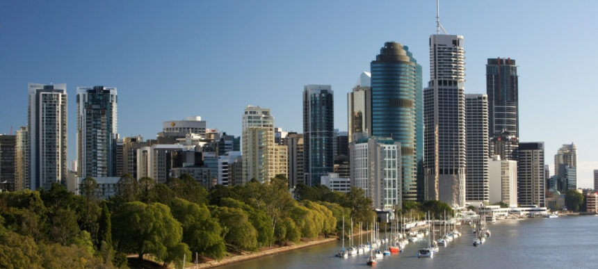 Brisbane city skyline viewed from the river, with high-rise buildings, parkland and moored boats along the waterfront.