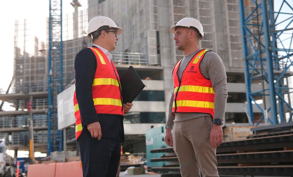 Two construction professionals in safety vests and hard hats speak at an active building site.