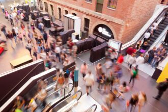A diverse crowd of people walking through a busy shopping mall, with stores visible in the background.