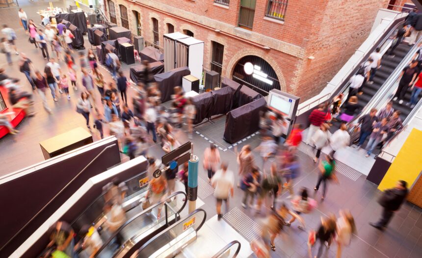 A diverse crowd of people walking through a busy shopping mall, with stores visible in the background.