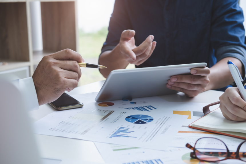 Three people sit at a table and analyse data on an ipad with various business documents showing graphs.