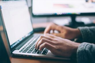 Person typing on a laptop keyboard with computer monitors in the background.
