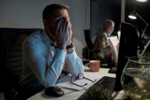 Small business owner working late at a desk, covering their face with their hands while reviewing paperwork and computer screens, illustrating business stress and workload pressures.