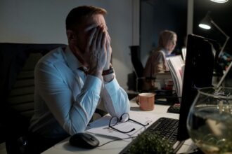 Small business owner working late at a desk, covering their face with their hands while reviewing paperwork and computer screens, illustrating business stress and workload pressures.