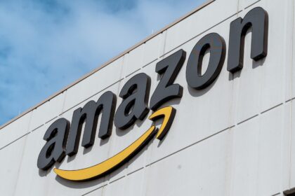 Close-up of the Amazon logo mounted on the exterior of a warehouse or fulfilment centre building against a blue sky.