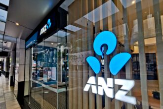 Exterior of an ANZ bank branch with illuminated blue ANZ logo signage displayed on the glass frontage. The entrance features automatic glass doors opening onto a modern lobby, with reflections of the surrounding city streetscape visible in the windows.
