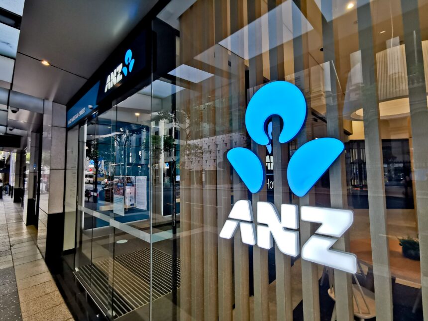 Exterior of an ANZ bank branch with illuminated blue ANZ logo signage displayed on the glass frontage. The entrance features automatic glass doors opening onto a modern lobby, with reflections of the surrounding city streetscape visible in the windows.