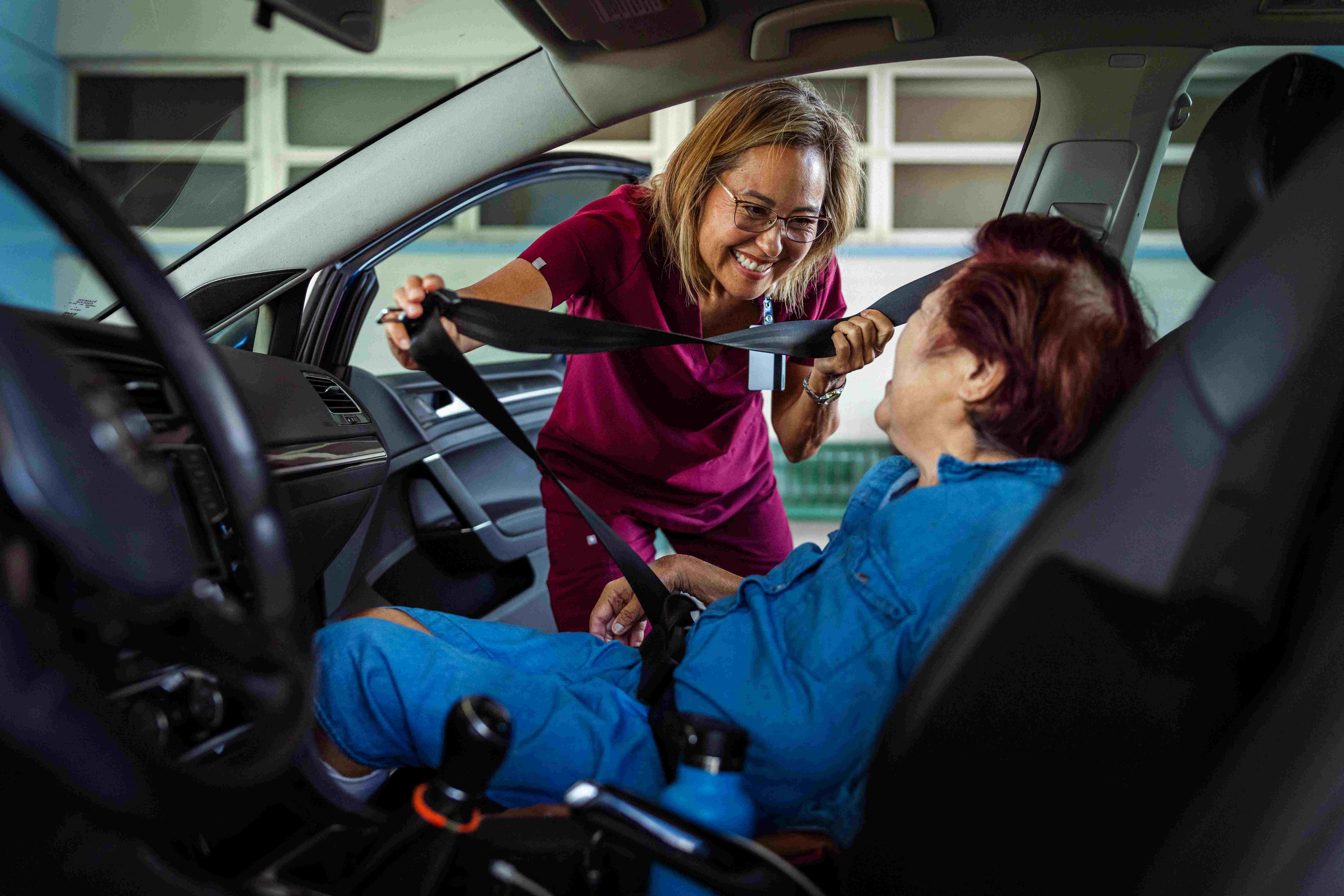 A healthcare worker helps an older person fasten their seatbelt while they are seated in the front passenger seat of a car, smiling as they prepare for a safe journey.