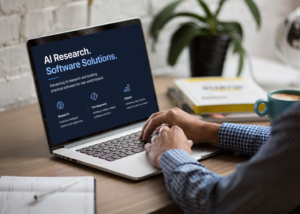 A person types on a laptop displaying an "AI Research. Software Solutions." webpage featuring sections on research, development and impact. The laptop sits on a desk beside a notebook and coffee cup, with a plant and books blurred in the background.