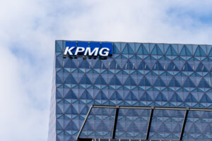 KPMG logo displayed on the upper facade of a modern glass office building against a cloudy blue sky.