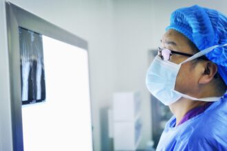 Healthcare professional wearing a surgical mask and protective clothing examining X-ray images on a lightbox in a clinical setting.