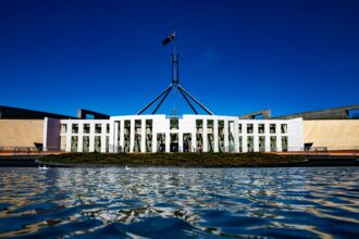 Exterior view of Australia's Parliament House in Canberra, with the Australian flag flying above the building and its reflection visible in the foreground water under a clear blue sky.