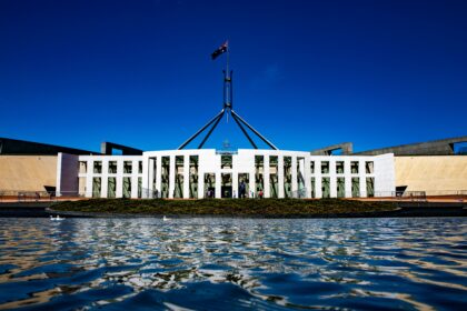 Exterior view of Australia's Parliament House in Canberra, with the Australian flag flying above the building and its reflection visible in the foreground water under a clear blue sky.