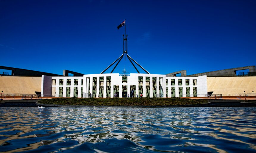 Exterior view of Australia's Parliament House in Canberra, with the Australian flag flying above the building and its reflection visible in the foreground water under a clear blue sky.