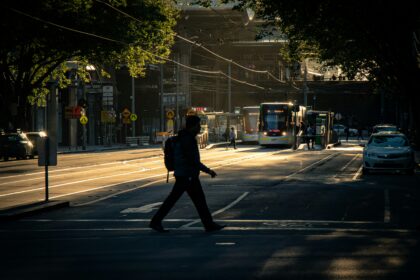 A pedestrian crosses a city street as trams approach in the background during late-afternoon sunlight.