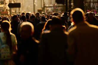 A large crowd of pedestrians walking along a busy city street in warm evening sunlight.