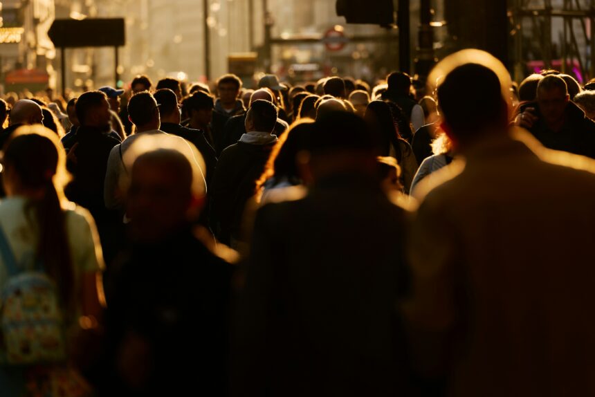 A large crowd of pedestrians walking along a busy city street in warm evening sunlight.