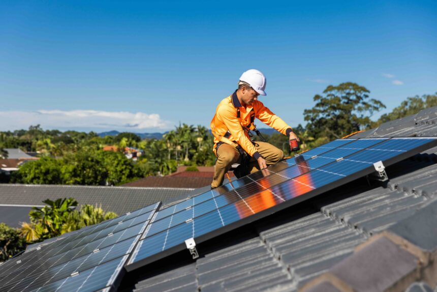 Technician wearing a hard hat, high-visibility clothing and safety harness installing rooftop solar panels on a residential home, using a power drill under clear blue skies.Technician wearing a hard hat, high-visibility clothing and safety harness installing rooftop solar panels on a residential home, using a power drill under clear blue skies.