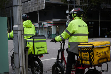 Pair of food delivery couriers on bikes, both wearing Uber Eats branded gear, sitting at an intersection in the city of Melbourne