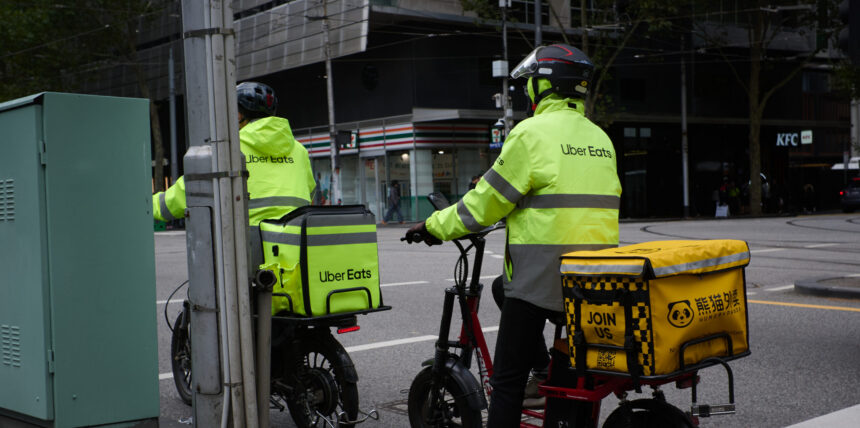Pair of food delivery couriers on bikes, both wearing Uber Eats branded gear, sitting at an intersection in the city of Melbourne