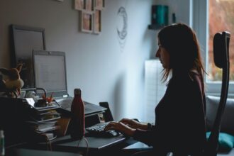 Woman working from home at a desk, typing on a keyboard beside a laptop.